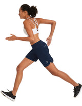 Woman in navy blue BALEAF 7" running shorts with white sports bra in motion. #color_Navy Blue