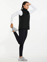 Black women's windproof softshell vest, white shirt, black leggings, gray sneakers. Woman stretches leg.