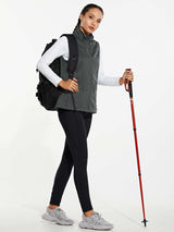 Woman in grey windproof softshell vest, white shirt, black leggings, grey sneakers, holding trekking pole and backpack.