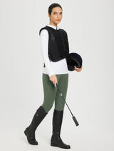 Woman in olive green riding breeches with full-seat silicone grip, black vest, and boots, holding helmet and whip. #color_Army Green