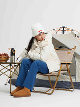 Woman in white puffer jacket and blue pants, seated with tent and camp table. Outdoor gear focus.