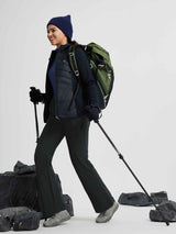 Woman hiking in black softshell pants, puffer vest, and navy beanie, with green backpack and trekking poles.