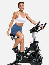 Woman in blue V-waist cycling shorts and white sports bra riding a stationary bike. #color_Navy Blue