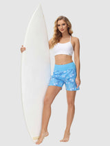 Woman in white sports bra and blue floral print 5" board shorts holding a white surfboard. #color_Print 2