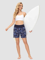 Woman in white sports bra and navy blue floral print swim shorts holding a white surfboard. #color_Print 1