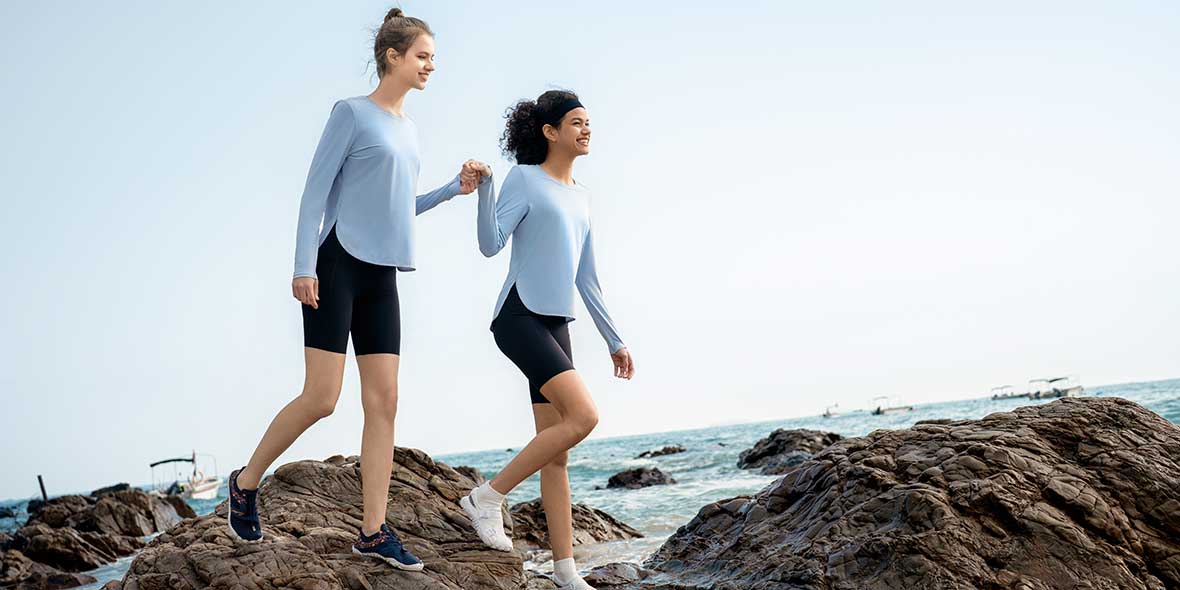 Two women in light blue long-sleeve sun shirts and black biker shorts walking along a rocky beach.