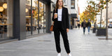 A woman wearing a black blazer and fleece-lined joggers holds a coffee cup while standing on a city sidewalk.