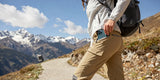 Realistic woman wearing women's hiking pants with deep pockets using phone on mountain trail.