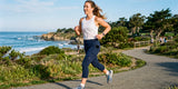 A woman in navy capri leggings with a phone in the side pocket runs along a coastal path