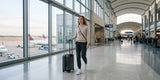 Woman wearing dark joggers, a light gray top, and white sneakers, pulling a carry-on suitcase through an airport terminal.
