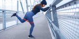 A woman in ankle-length leggings performing a resistance-band glute kickback against a bridge railing.