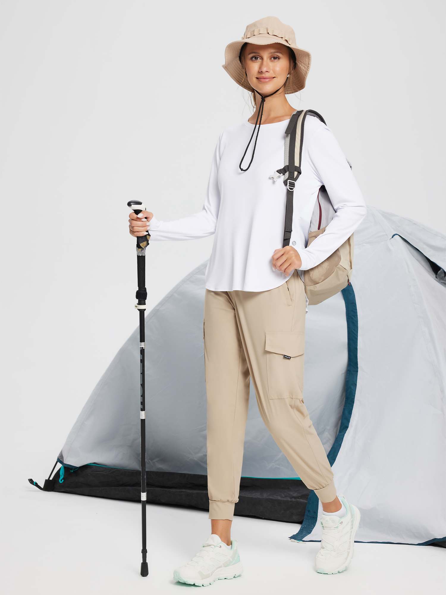 Woman in white long sleeve shirt, khaki cargo pants, sun hat, holding trekking pole near tent. #color_White