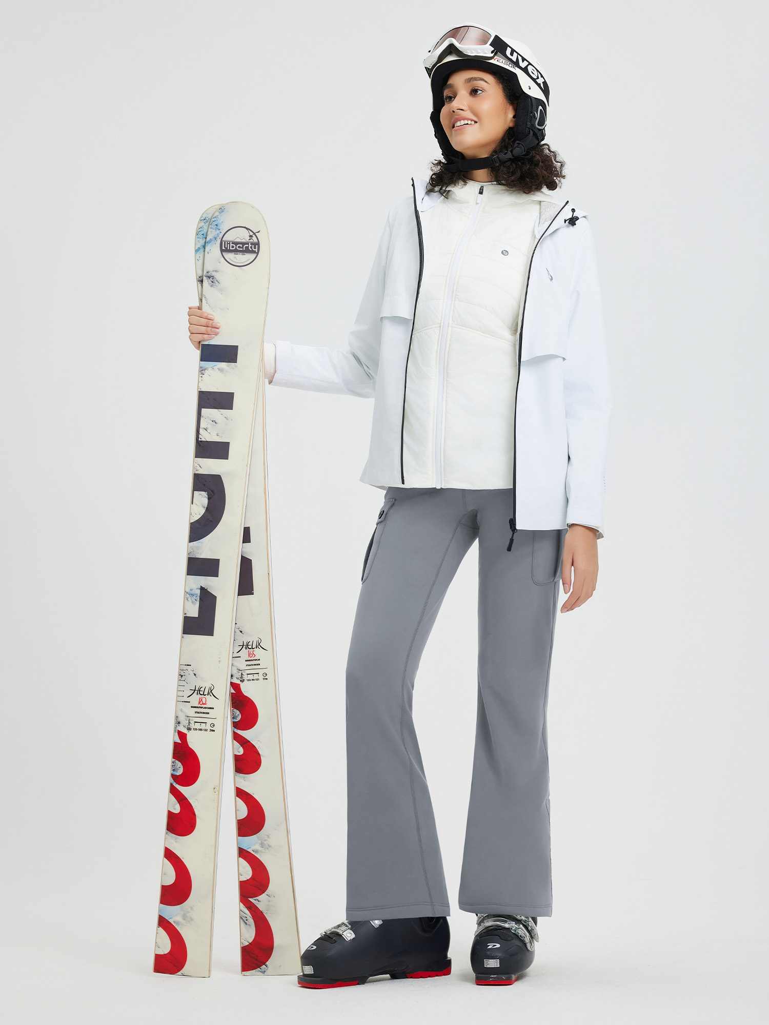 Woman in grey flared outdoor pants, white vest, blue jacket, holds skis, wears ski gear. #color_Dark Gray