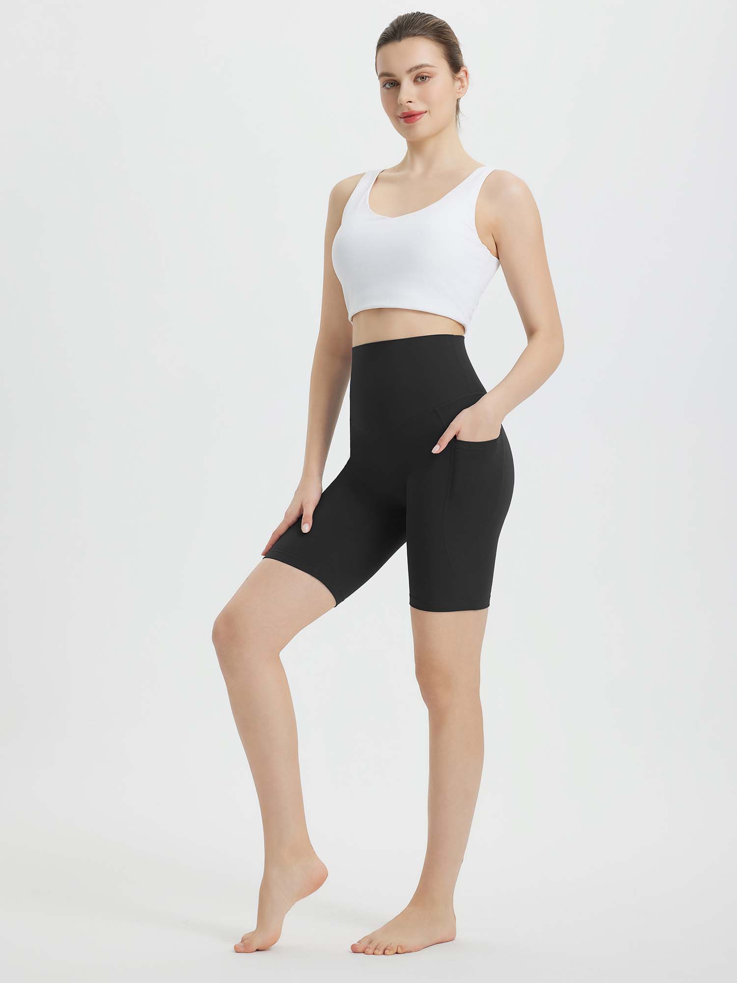 Woman in black 8-inch biker shorts with high waist and side pockets, paired with a white sports bra. #color_Black