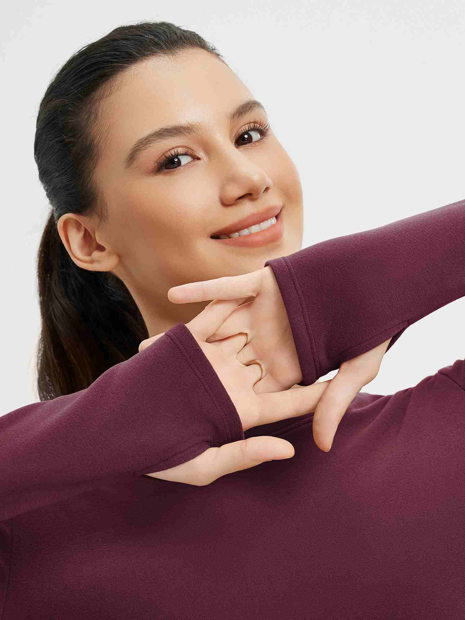 Woman wearing a burgundy side-ruched long-sleeve yoga top with thumbholes, smiling and stretching her arms. #Color_Wine Red