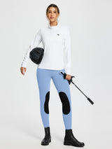 Light blue knit skinny breeches for horse riding with black knee patches. Woman holds helmet and crop. #color_Light Blue