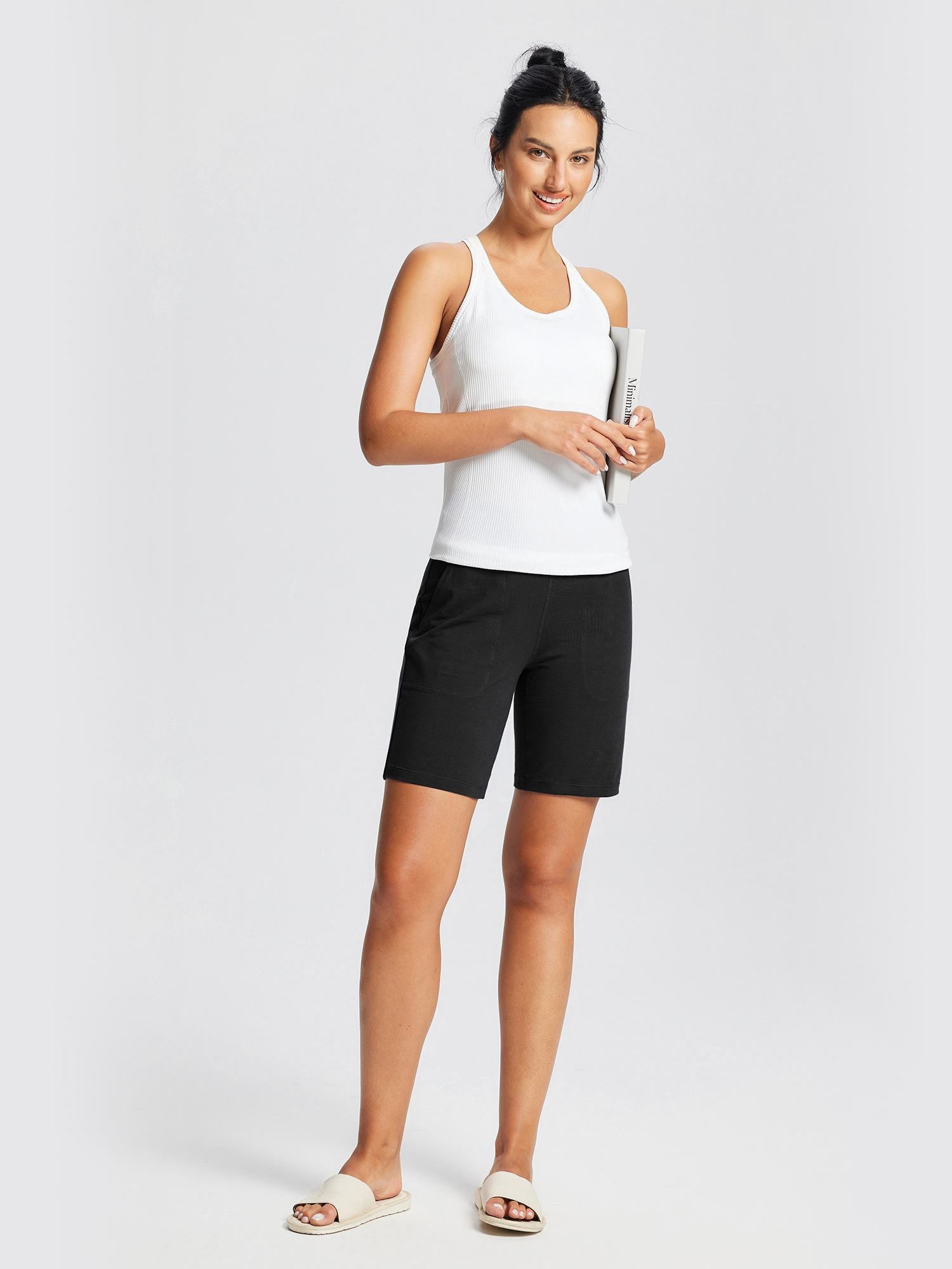 Woman wearing black 8" knit waistband yoga shorts and white ribbed tank top, holding a book. #color_Black