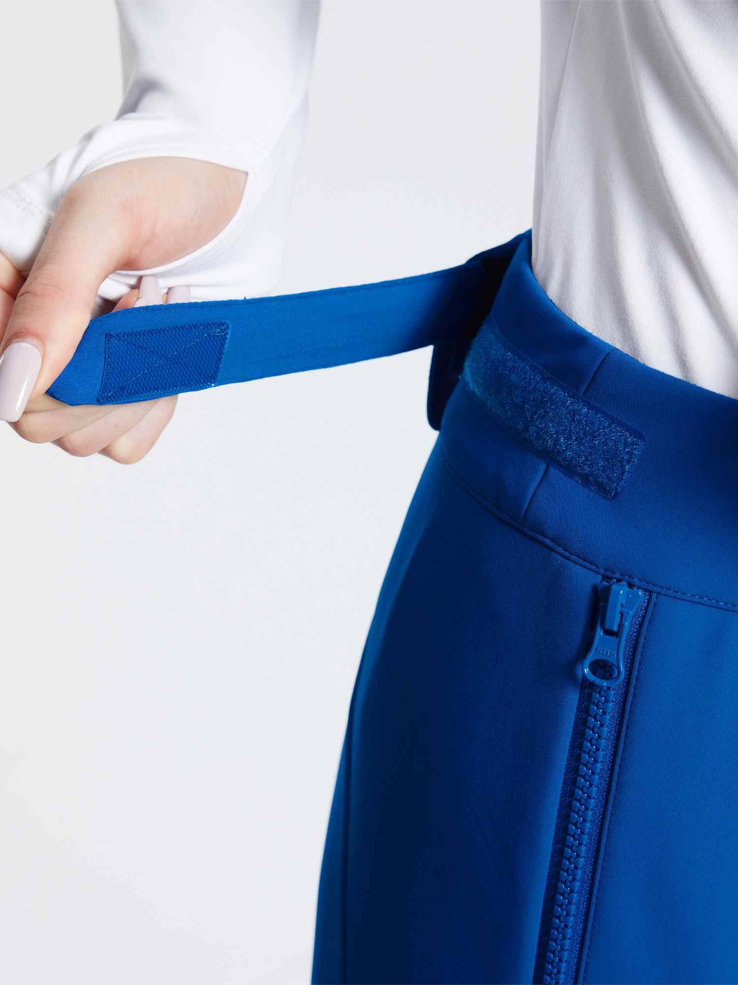 Woman adjusting velcro waist strap on blue stretch knit slim-fit ski pants with zippered pocket. #color_Quartz Blue
