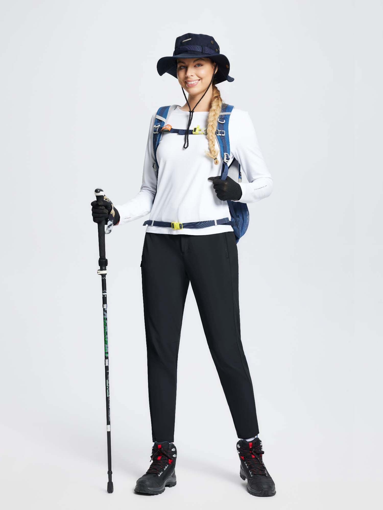 Woman in black slim-fit ski pants, white top, backpack, hat, and boots holding trekking pole. #color_Black