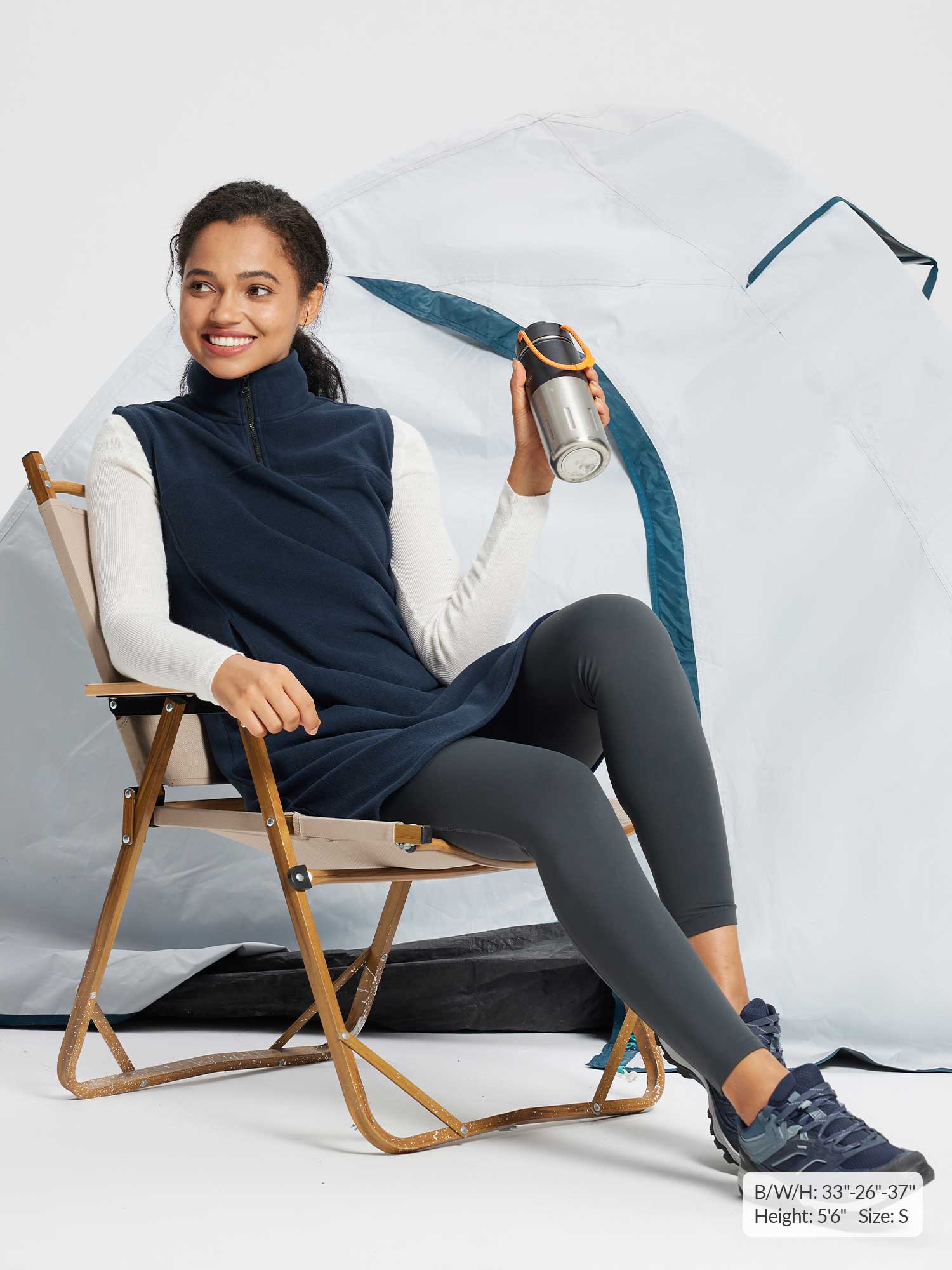 Navy sleeveless fleece thermal dress, quarter-zip, worn over white long-sleeve shirt, grey leggings, and sneakers. Model holding water bottle by tent.