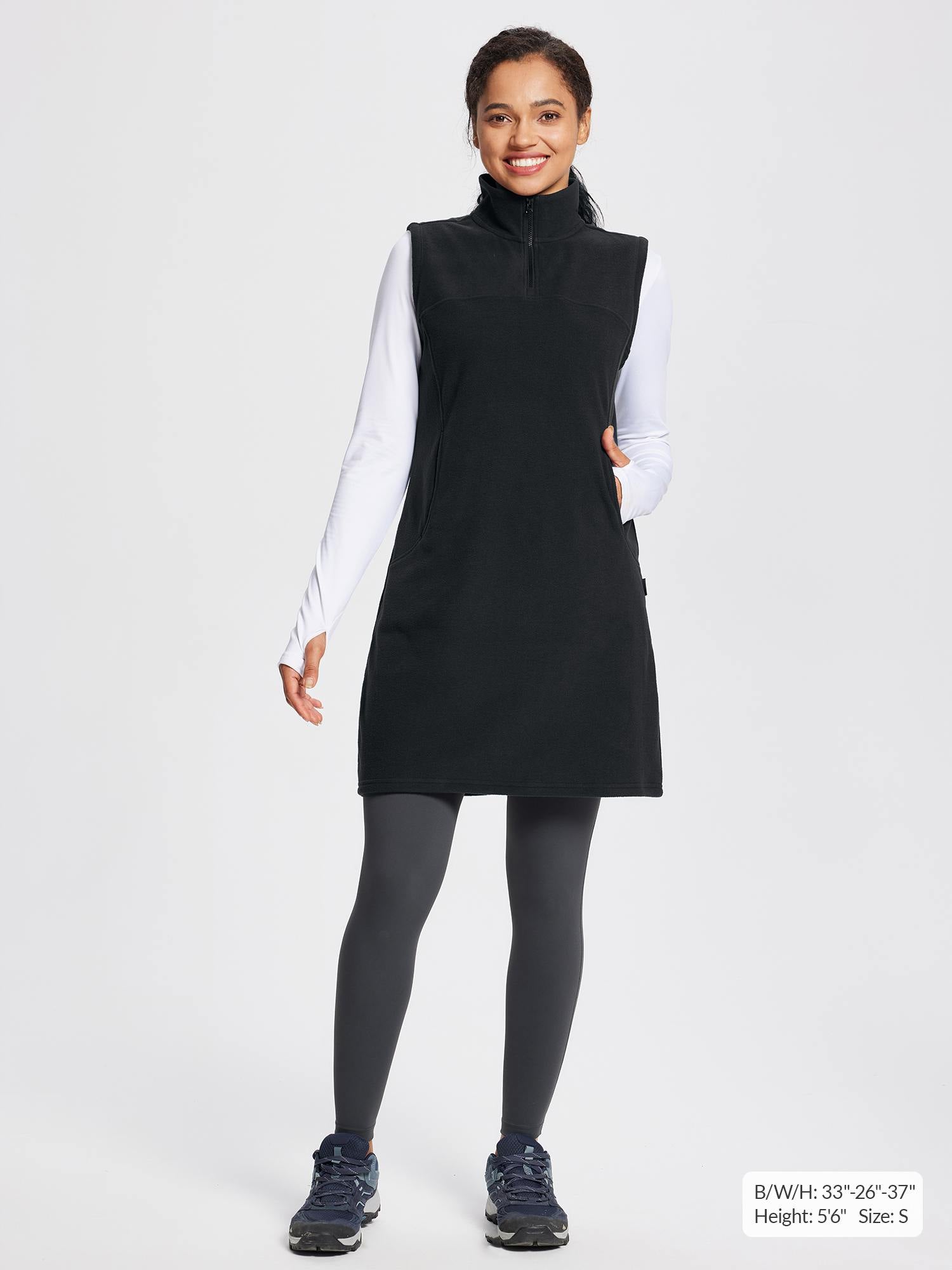 Black sleeveless fleece thermal dress with quarter-zip and pockets, worn over white long-sleeve shirt and grey leggings.